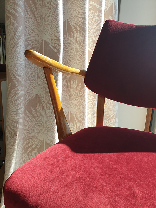 Close-up of a wooden chair with burgundy fabric upholstery in front of a beige curtain with a leaf pattern.