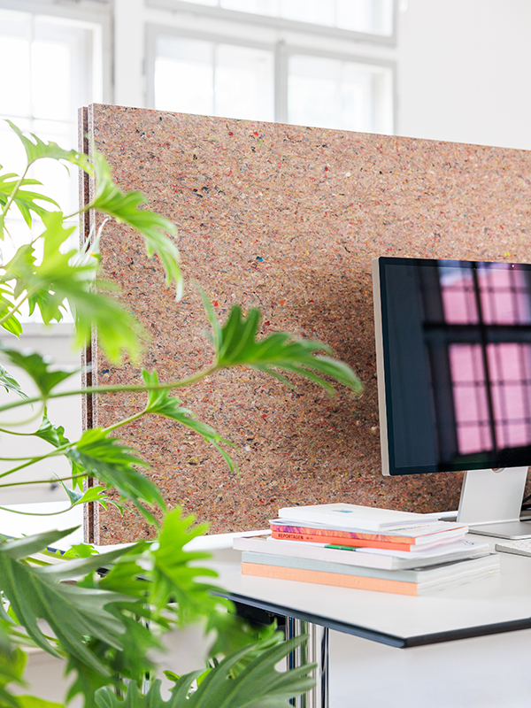 Office desk with a computer monitor, stacked books, and a green leafy plant in front of a large textured cork or recycled material panel.