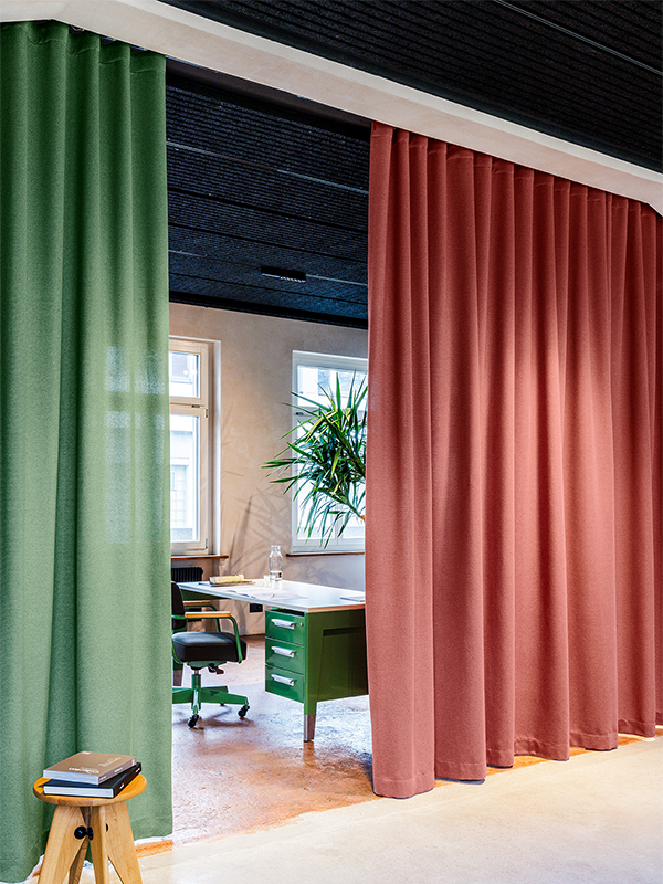 Office space with green and rust-colored floor-to-ceiling curtains partially open, showing a desk, chair, windows, and a large indoor plant.