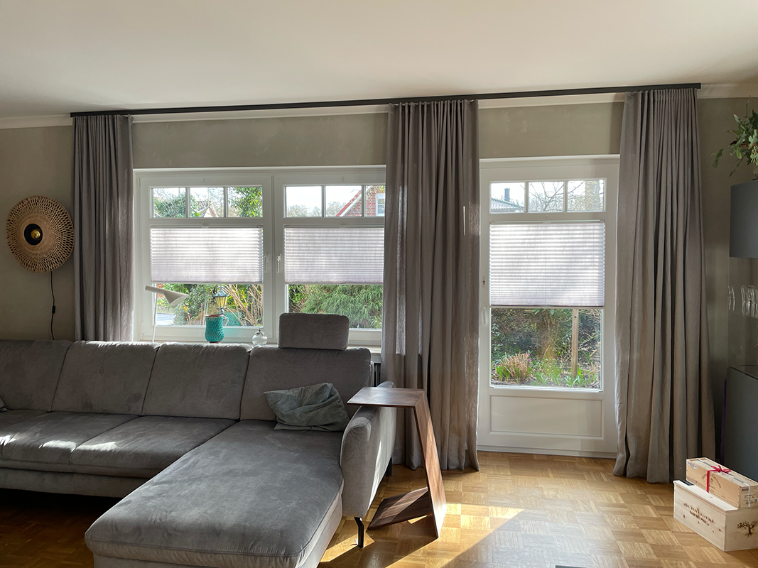 Living room with gray sectional sofa, wooden side table, large windows with gray curtains and white blinds partially closed.