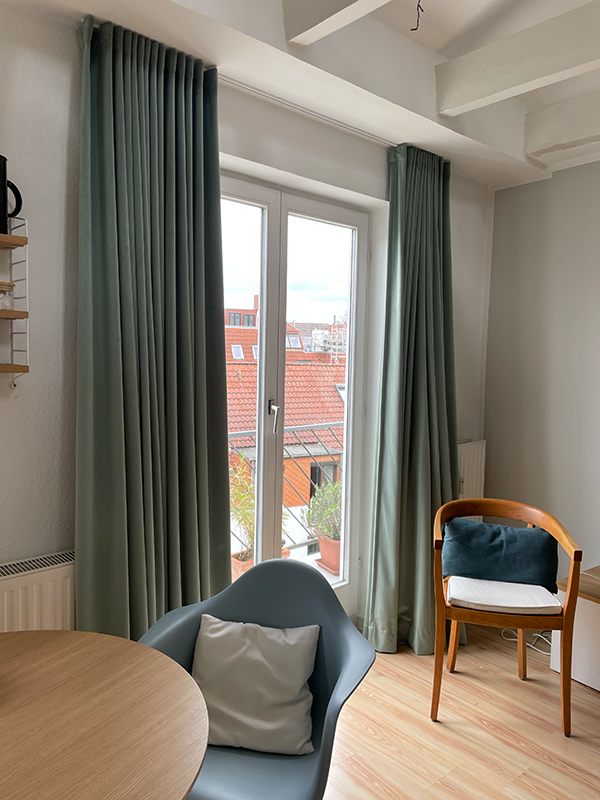 Room interior with light wood flooring, large window with green curtains, a round wooden table, a blue armchair with a white pillow, and a wooden chair with a dark blue cushion.