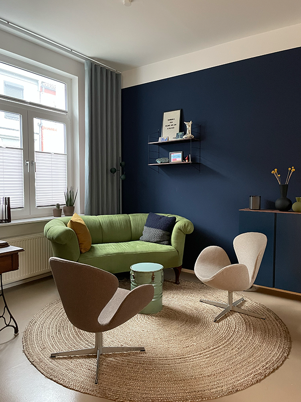 Modern living room with a green sofa, two beige swivel chairs, round woven rug, small green round table, and navy blue accent wall with shelves and decor.