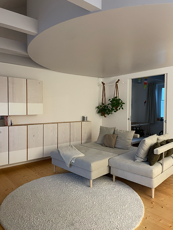 Modern living room with a light gray sectional sofa, beige and brown pillows, a round gray rug on wooden floor, white wall cabinets, and two hanging plants near a doorway.