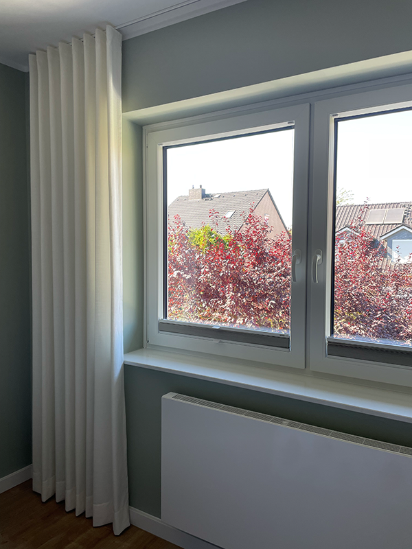 White curtain and radiator beneath a double-pane window with a view of houses and red-leaved bushes outside.