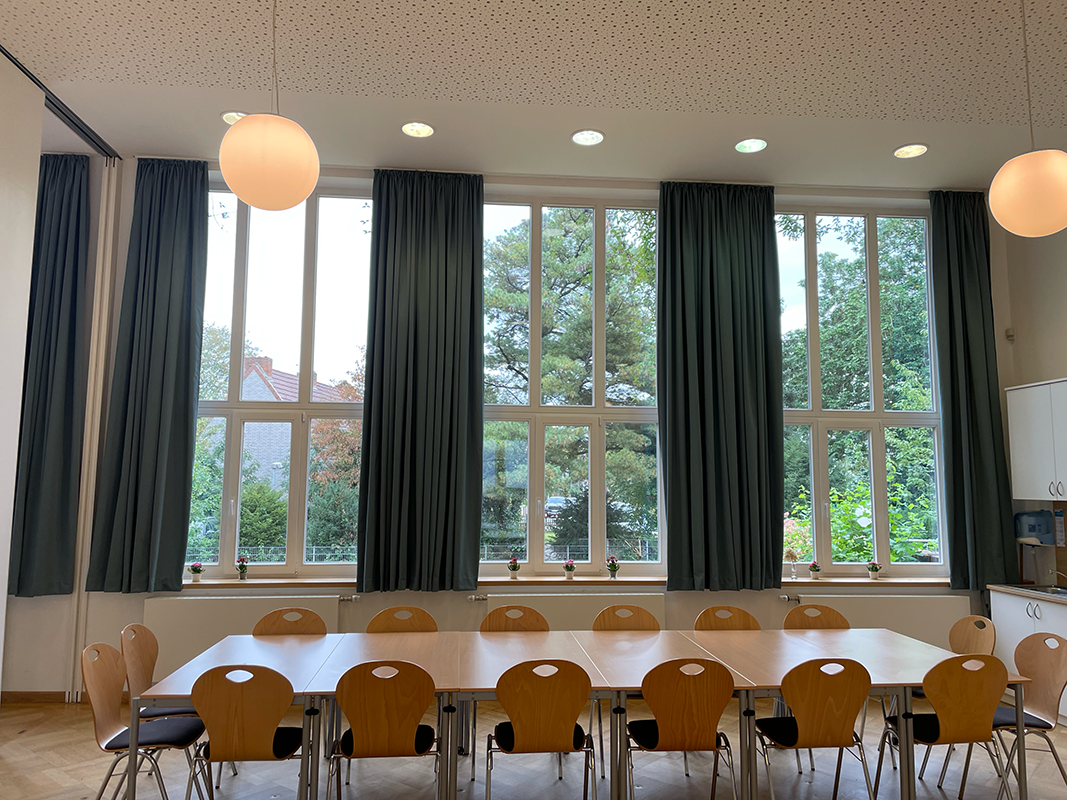 Conference room with a long table surrounded by wooden chairs facing large windows with green curtains and a view of trees outside.