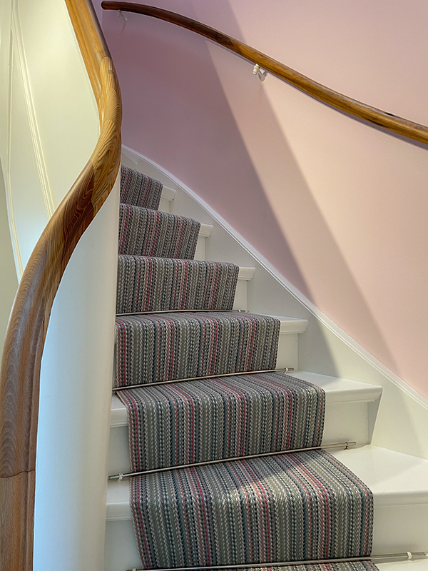 Curved white staircase with a striped gray and pink carpet runner secured by metal rods, wooden handrails, and a pale pink wall.