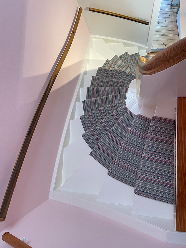 Curved white staircase with multi-colored striped stair treads and wooden handrails, viewed from above.