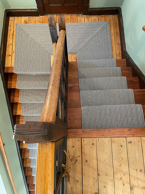Top view of wooden staircase with patterned black and white runners on each step, a wooden handrail, and surrounding wooden floor flooring.