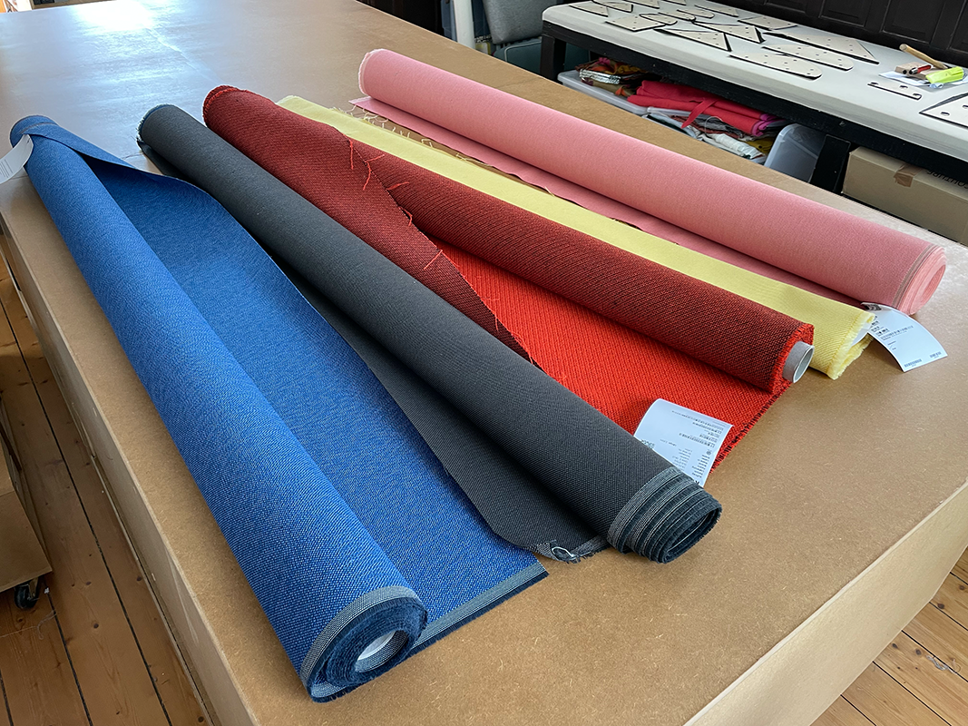 Five rolls of fabric in blue, dark gray, red, yellow, and pink laid out on a wooden table in a workshop.