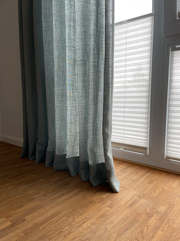 Light blue textured curtains partially covering a window with closed white blinds and a wooden floor below.