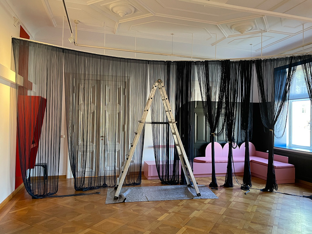 Interior room with wooden parquet floor, black sheer curtains hanging from ceiling, a silver step ladder on a gray mat, a pink sectional sofa, and a large window letting in natural light.