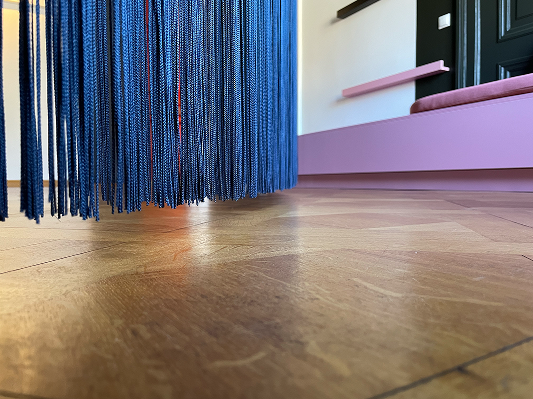 Close-up of blue hanging strands near a wooden floor with a pink and black wall shelf in the background.