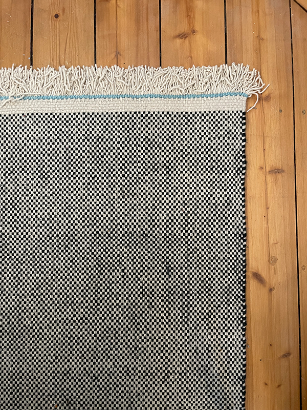 Close-up of a black and white checkered woven rug with white fringe and a thin blue stripe on a wooden floor.