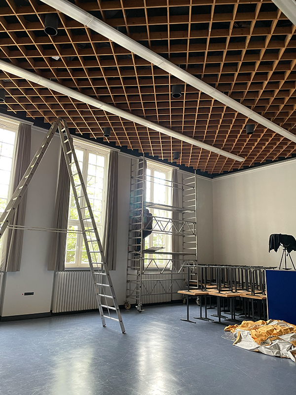 Indoor room with a high wooden grid ceiling, large windows, scaffolding with a person working, a tall ladder, stacked tables, and a pile of fabric on the floor.