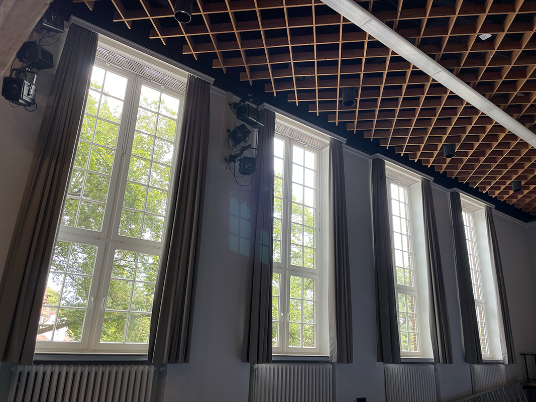 Four tall, white-framed windows with dark curtains in a room with a wooden grid ceiling and wall-mounted stage lights.