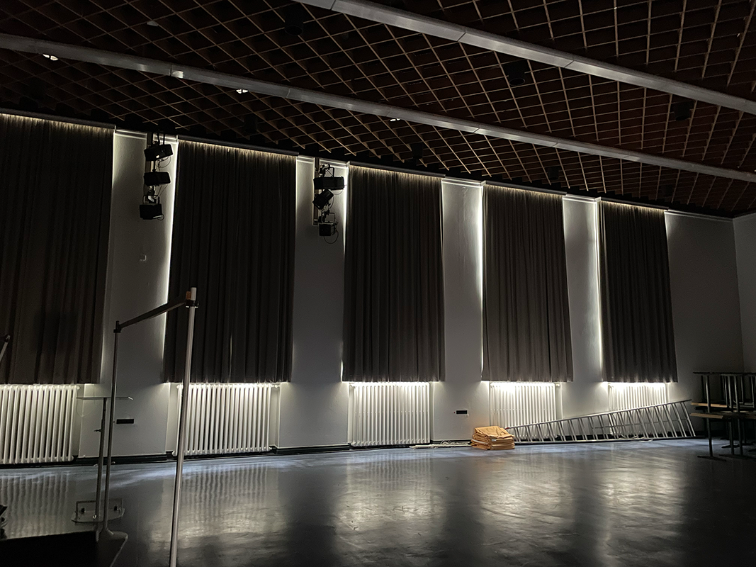 Dimly lit room with several large curtains partially covering windows, stage lights mounted on the wall, and a metal ladder leaning against the wall near folded yellow fabric.