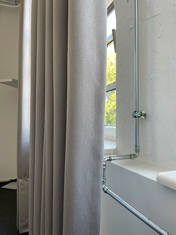Light gray pleated curtain next to a white wall with exposed metal piping and a window showing green foliage outside.