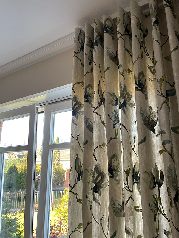 Beige floral-patterned curtain partially covering a sunlit window with a garden view outside.