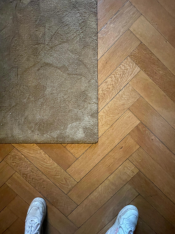 White sneakers on wooden herringbone parquet floor partially covered by beige carpet corner.