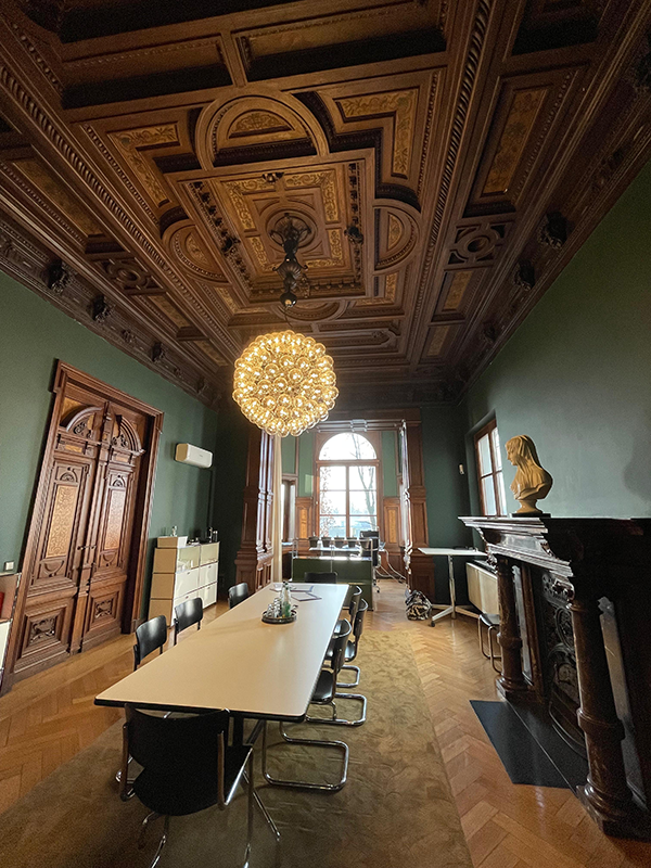 Conference room with an ornate wooden ceiling, large wooden doors, a modern light fixture, long white table with black chairs, and a white bust on a dark wooden mantel.