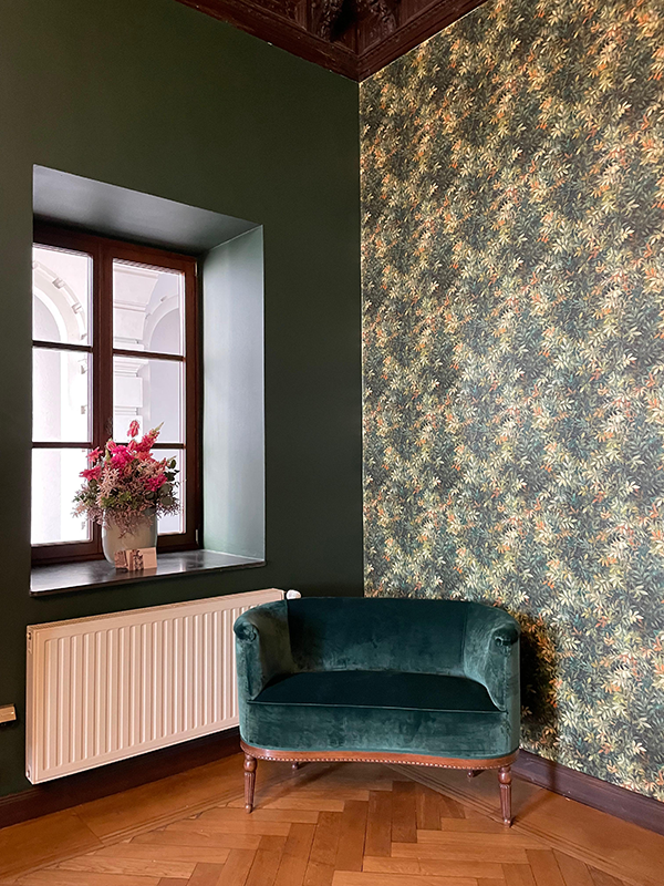 Green velvet corner armchair next to a window with floral wallpaper and a vase of pink flowers on the windowsill.