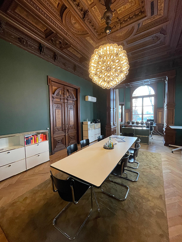 Elegant meeting room with a long white table surrounded by black chairs, ornate wooden ceiling, and large chandelier.
