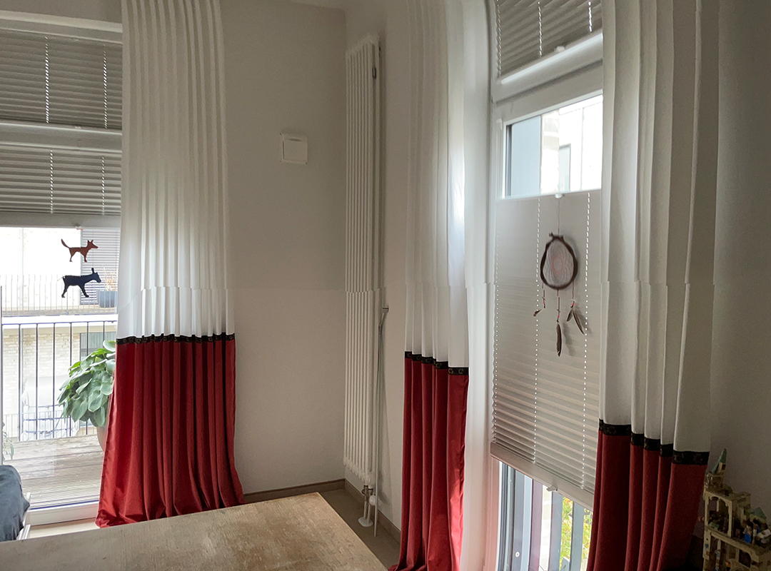 Interior corner with two windows featuring white and red curtains, pleated blinds, a dreamcatcher, and small decorative dog figures on one window.