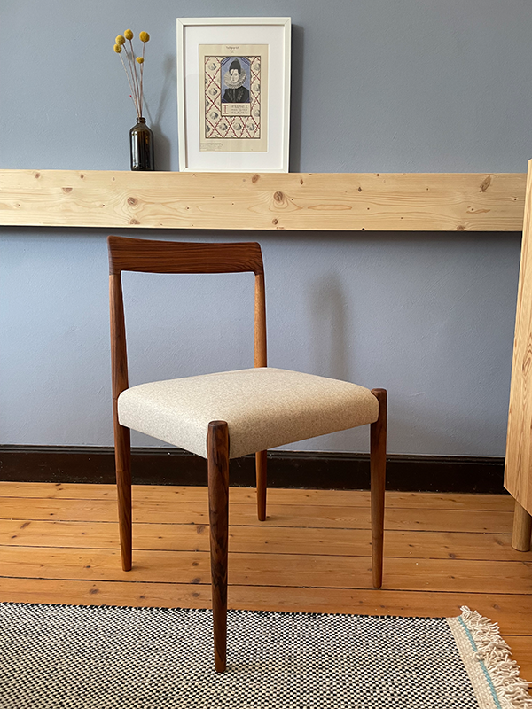 Wooden chair with beige upholstered seat on wooden floor in front of gray wall with framed artwork and small vase with yellow flowers on a wooden shelf.