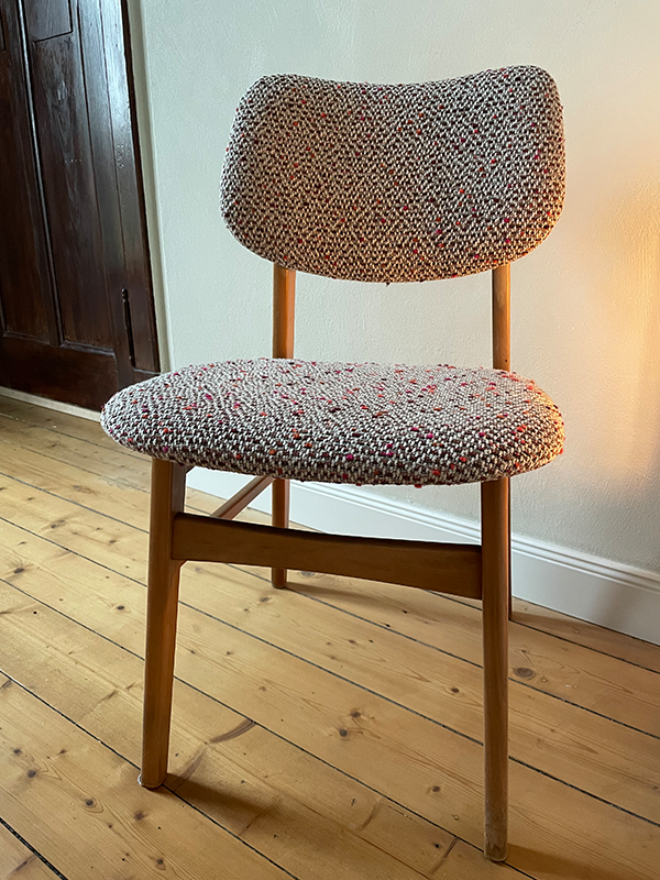 Wood-framed chair with curved backrest and seat covered in textured multicolored fabric on wooden floor against a white wall.