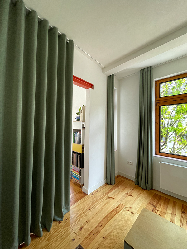 Room corner with light wooden floor, green curtains hanging near a window and doorway leading to a room with shelves filled with books and records.