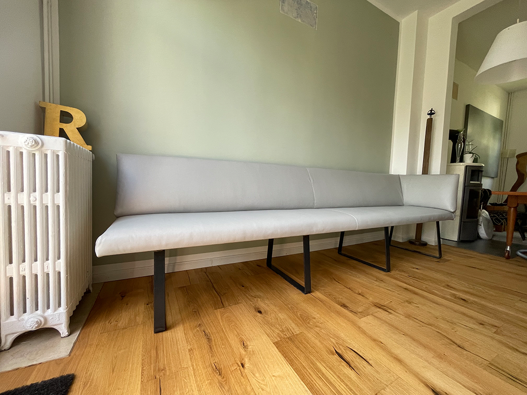 Modern light gray upholstered bench with black metal legs on wooden floor against a pale green wall next to a white radiator with a yellow letter R on it.