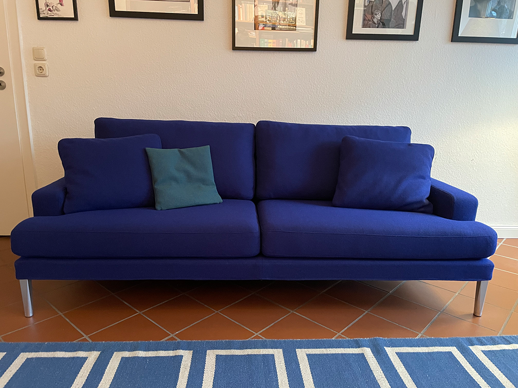 Modern dark blue sofa with metal legs and three cushions, two matching blue and one teal, on a tiled floor with a blue and white patterned rug in front.