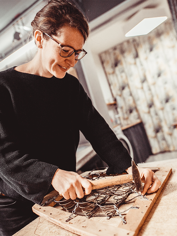 Person wearing glasses and a black sweater working with a hammer on a wooden frame with metal springs indoors.